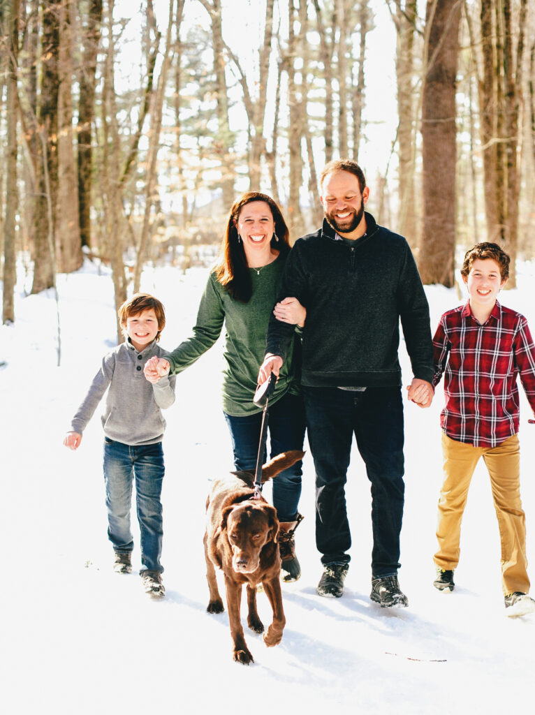 a smiling family walks through snowy woods with their dog